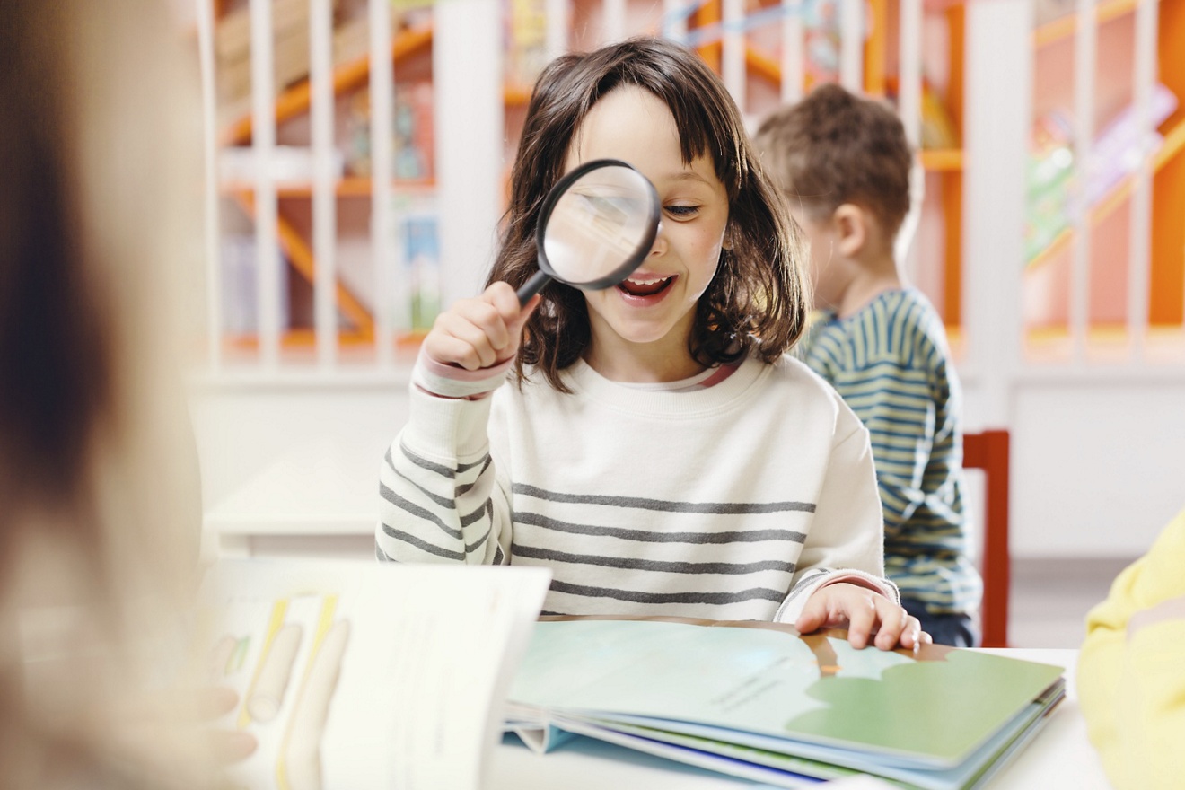 bookshop, library, indoors, kids, sitting, activity, people, casual, stripy, smiling, mid-length hair, brown hair, illustrated book, desk, holding, magnifying glass, looking through, classroom, concentration, toothy smile, happiness