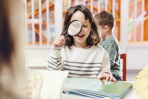 bookshop, library, indoors, kids, sitting, activity, people, casual, stripy, smiling, mid-length hair, brown hair, illustrated book, desk, holding, magnifying glass, looking through, classroom, concentration, toothy smile, happiness