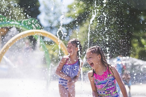 A group of young children frolic and run in a playground splash pad. It is a sunny, perfect day for getting wet and playing hard! Two young girls in swimsuits are running through the sprinklers and yelling and laughing as the water droplets cascade all around them.