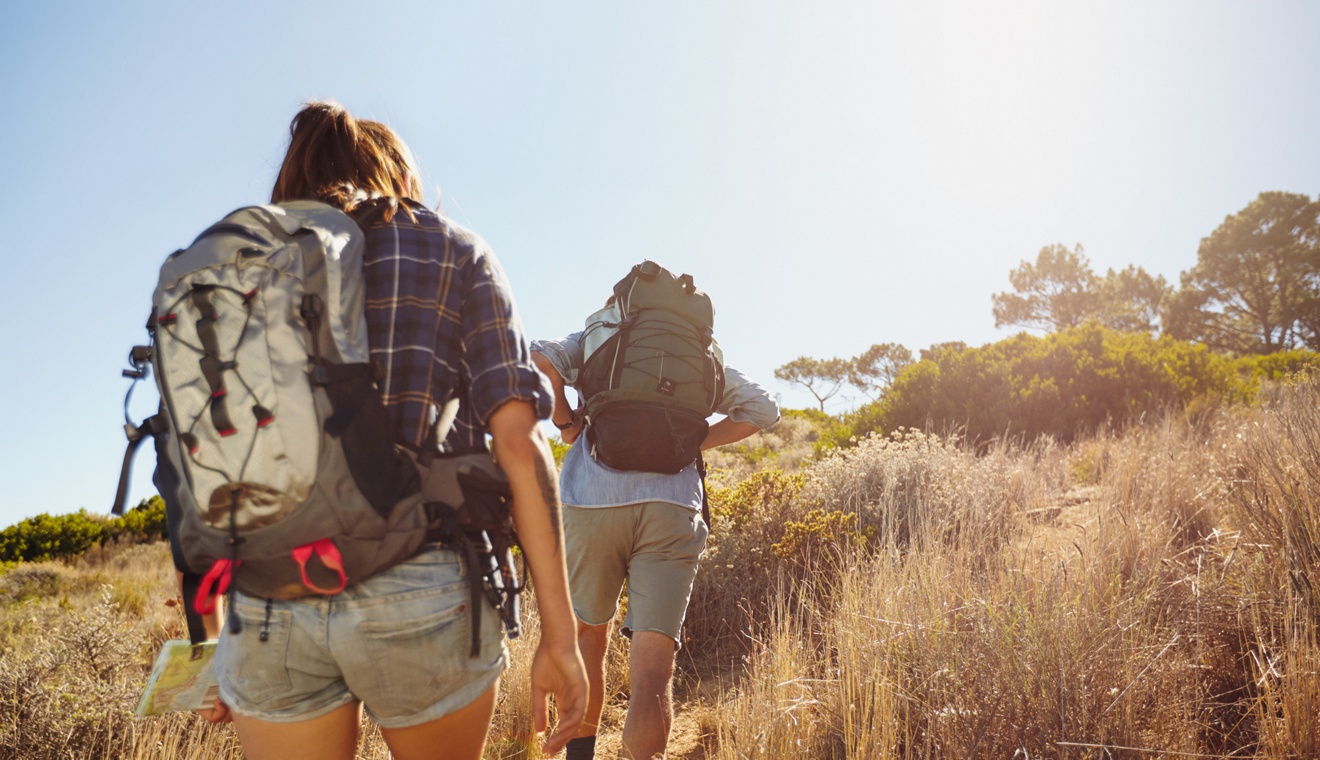 Rear view image of two young people carrying backpacks walking through mountain trial on summer day. Man and woman hiking on bright sunny day.