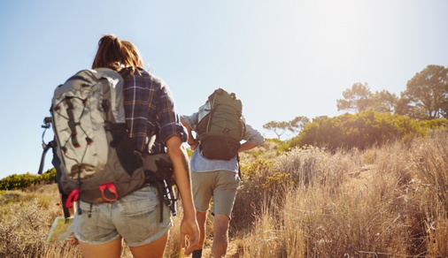 Rear view image of two young people carrying backpacks walking through mountain trial on summer day. Man and woman hiking on bright sunny day.