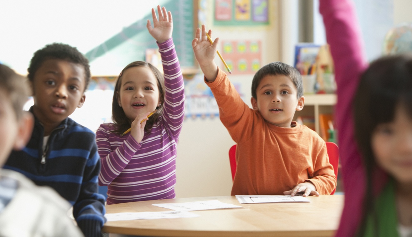 Image of first grade students raising their hands in a classroom.
