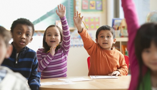 Image of first grade students raising their hands in a classroom.