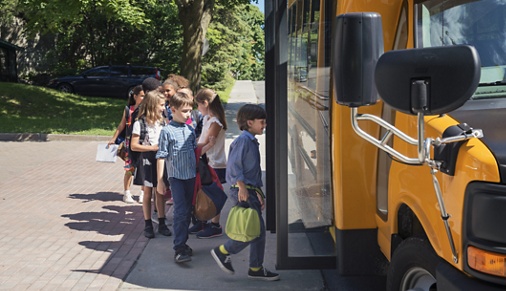 Group of elementary school kids getting in a yellow school bus. Multi-ethnic group, boys and girls age 8-9, with books, backpack and lunch boxes. A little boy is leading the pack, ready to get in the bus, the door is open. Horizontal outdoors shot with copy space. This was taken in Quebec, Canada.