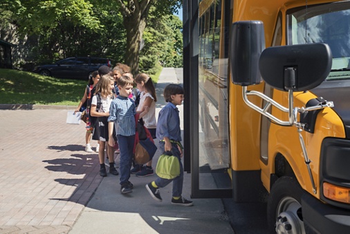 Group of elementary school kids getting in a yellow school bus. Multi-ethnic group, boys and girls age 8-9, with books, backpack and lunch boxes. A little boy is leading the pack, ready to get in the bus, the door is open. Horizontal outdoors shot with copy space. This was taken in Quebec, Canada.